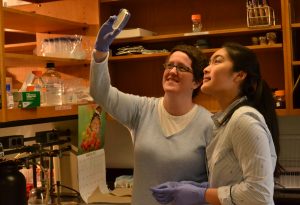 Dr. Erin Green holds petri dish up to light as her and student look through it.